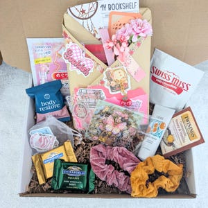 May include: A gift box filled with various items, including a Swiss Miss hot chocolate mix, Ghirardelli chocolate squares, a body restore packet, and a variety of stickers. The box also contains a pink scrunchie, a yellow scrunchie, and a small tube of hand cream.