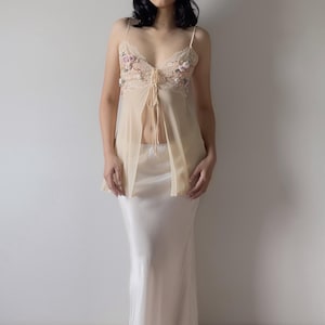 Könnte beinhalten: Cremefarbenes Dessous-Set. Das transparente Oberteil mit offenem Schnitt hat Blumenstickereien und Spaghettiträger mit Bindeband. Der passende Rock ist aus Satin und bodenlang. Ein zartes, elegantes Design.
