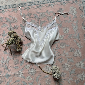 Puede incluir: Camisola de satén blanco con delicados ribetes de encaje y tirantes finos. La camisola presenta un escote en V con un panel transparente de lunares. La prenda se exhibe sobre una superficie con estampado floral rosa.