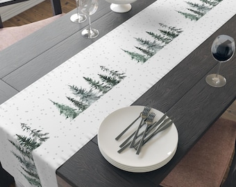 Puede incluir: Un camino de mesa blanco con un diseño de acuarela de pinos verdes y copos de nieve blancos. El camino de mesa está colocado sobre una mesa de madera oscura con un cuenco de manzanas, dos copas de vino y una pila de platos blancos.