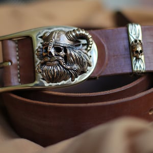 May include: A brown leather belt with a brass buckle featuring a detailed skull design with a horned helmet. The buckle is oval-shaped and has a textured surface. The belt has a second brass accent with a smaller skull design.