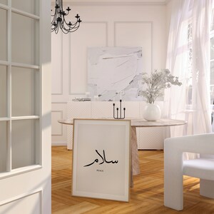 May include: A framed print with the Arabic word for peace, "Salam", in black calligraphy on a cream background. The word "Peace" is written in English below the Arabic text. The print is hanging on a white wall in a modern living room.