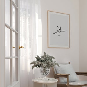 May include: A white framed print with the Arabic word for peace, "Salam", in black calligraphy. The word "Peace" is written in English below the Arabic text. The print is hanging on a light pink wall, with a white sheer curtain and a wooden door in the background. A vase with white flowers sits on a white side table.