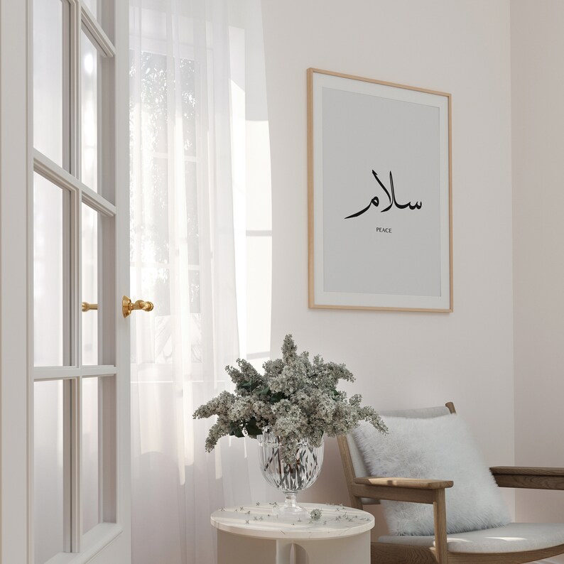 May include: A white framed print with the Arabic word for peace, "Salam", in black calligraphy. The word "Peace" is written in English below the Arabic text. The print is hanging on a light pink wall, with a white sheer curtain and a wooden door in the background. A vase with white flowers sits on a white side table.