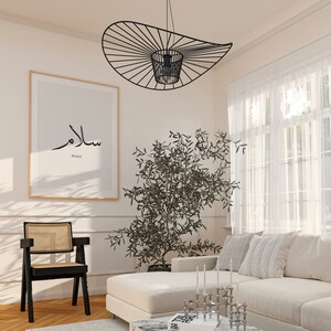 May include: A white couch with black and white throw pillows in a living room with a black metal light fixture, a potted olive tree, and a framed print with the word "Salam" in Arabic script and the word "Peace" in English.