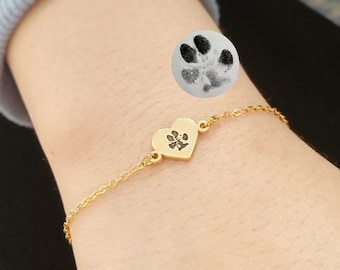 May include: A gold heart-shaped charm bracelet with a paw print engraved on the charm. The bracelet is on a person's wrist.