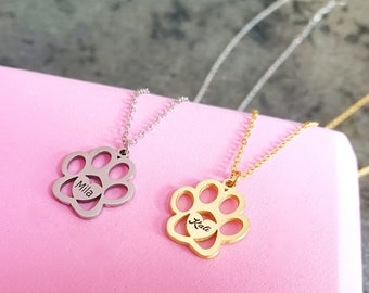 May include: Two silver and gold necklaces with paw print pendants. The silver necklace has the name "Mila" engraved on the pendant. The gold necklace has the name "Kelli" engraved on the pendant.
