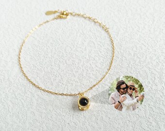 May include: A gold chain bracelet with a small round pendant that has a black photo inside. The photo shows a couple smiling and taking a selfie.