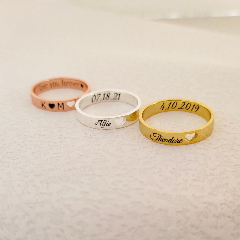 May include: Three personalized rings with heart cutouts. The silver ring says "07.18.21 Alfie", the gold ring says "4.10.2019 Theodore", and the rose gold ring says "love you forever K♡M".