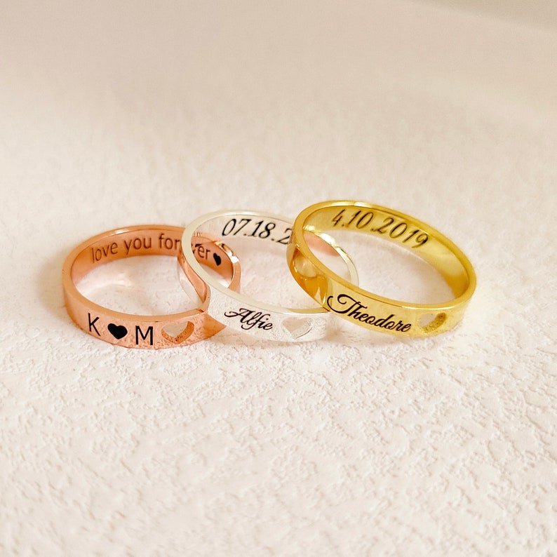 May include: Three personalized rings with heart cutouts. The gold ring says "Theodore" and "4.10.2019". The silver ring says "Alfie" and "07.18.20". The rose gold ring says "love you forever" and "K M".