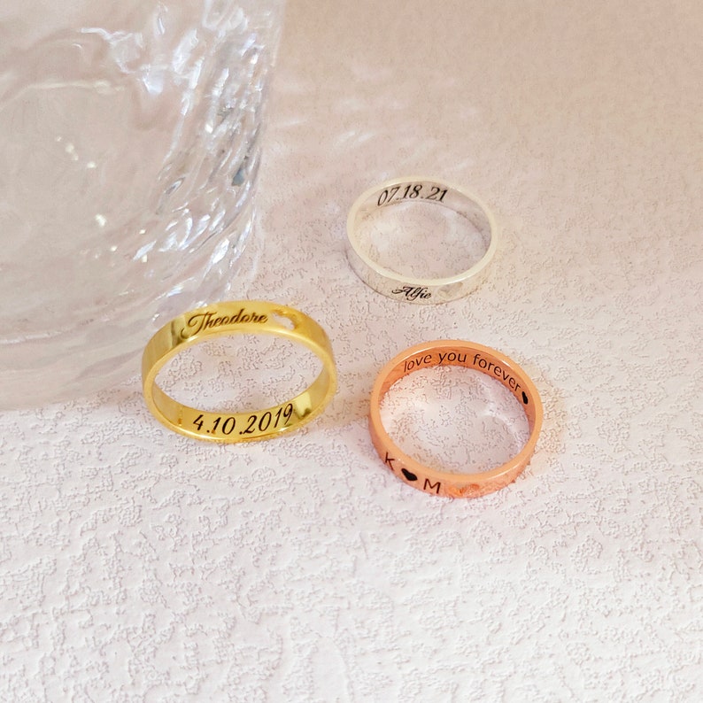 May include: Three personalized rings with engraved text. The gold ring says "Theodore" and "4.10.2019". The rose gold ring says "love you forever" and "K & M". The silver ring says "Alfie" and "07.18.21".