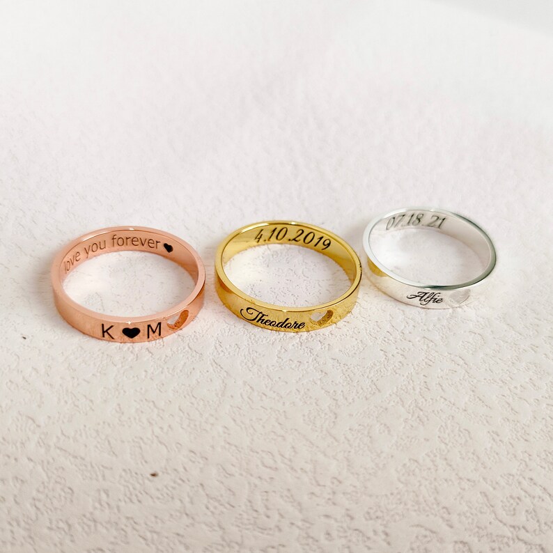 May include: Three personalized rings with engraved text. The rose gold ring says "love you forever" with the initials "K" and "M" separated by a heart. The gold ring says "Theodore" with the date "4.10.2019". The silver ring says "Alfie" with the date "07.18.21".
