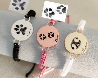 Puede incluir: Tres pulseras personalizadas con dijes de huella de pata y los nombres "Molly", "Oscar" y "Callie" grabados en los dijes. Las pulseras están hechas con cordones negros, rosas y blancos.