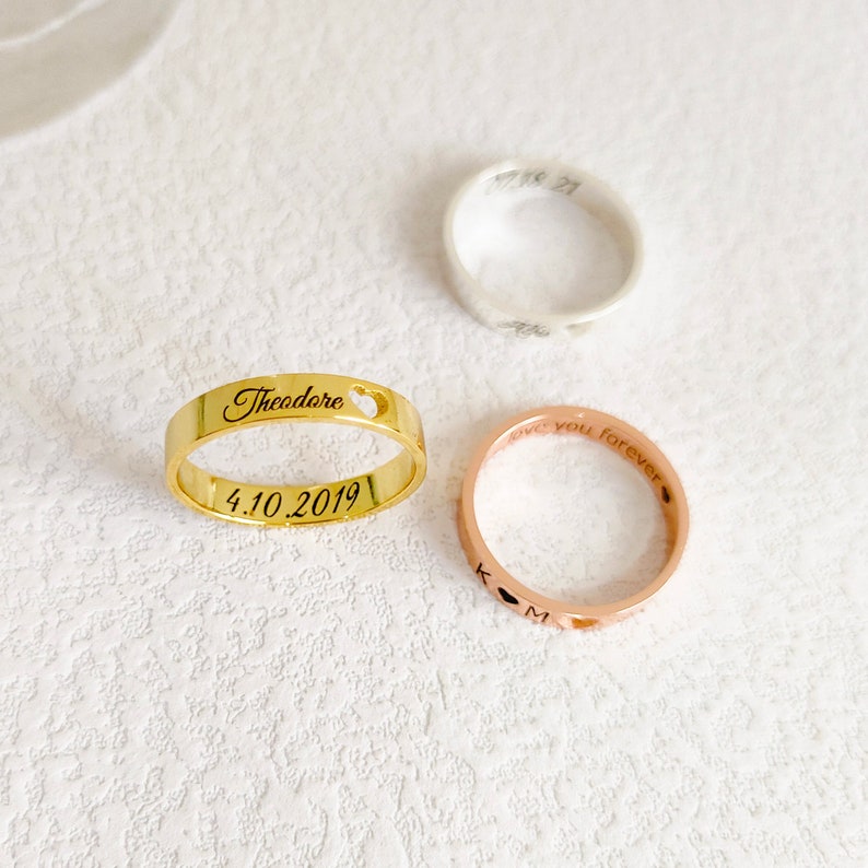 May include: Three gold, silver, and rose gold rings with personalized engravings. The gold ring says "Theodore" with a heart cutout and the date "4.10.2019". The rose gold ring says "love you forever" and has a heart cutout. The silver ring has a heart cutout and the date "11.18.21".