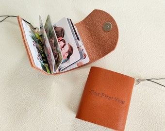 May include: A brown leather photo album with a strap and a metal clasp. The album is open to reveal several photos of a couple. The album is labeled "Our First Year".