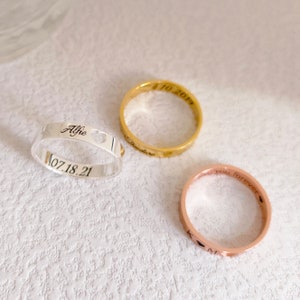 May include: Three silver, gold, and rose gold rings with engraved text. The silver ring says "Alfie 07.18.21" and has a heart cut out. The gold ring says "11.10.2019" and "Forever." The rose gold ring says "Love you forever" and has a heart cut out.