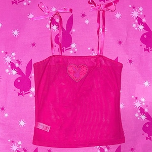 Puede incluir: Un top de tirantes de malla rosa con un logotipo de conejito Playboy rosa en forma de corazón. El top tiene tirantes finos y un lazo rosa en cada tirante.