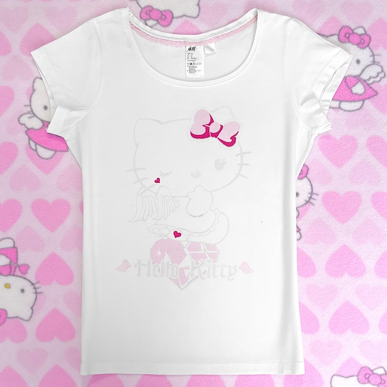 May include: White t-shirt with a Hello Kitty design, including a pink bow, heart, and the words "Hello Kitty". It has a scoop neck with pink trim and short sleeves. The background shows a pink heart pattern.