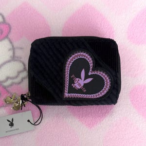 May include: Black corduroy wallet with a heart-shaped patch featuring the Playboy logo in purple. The wallet has a zipper closure and a small tag with the Playboy logo and the word "accessories".