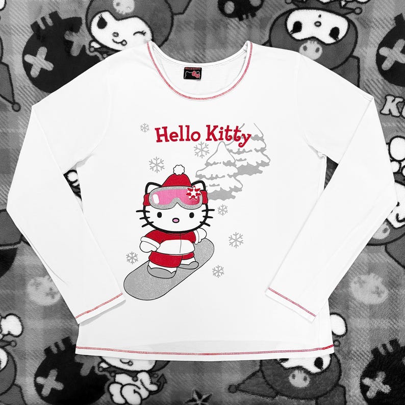 Pu&ograve; includere: Maglietta bianca a maniche lunghe con rifiniture rosse, con una grafica di Hello Kitty che fa snowboard. Il disegno mostra Hello Kitty con un abito rosso, occhiali rosa e un cappello rosso. Le parole "Hello Kitty" sono sopra.