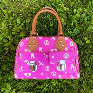 May include: A pink handbag with a brown handle and white bunny rabbit designs. The bag has two front pockets with silver clasps.