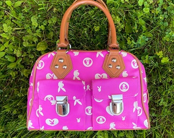 May include: A pink handbag with a brown handle and white bunny rabbit designs. The bag has two front pockets with silver clasps.