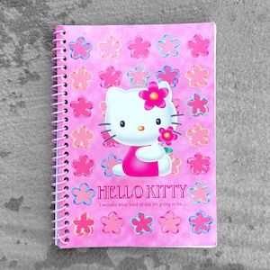 May include: Pink spiral-bound notebook featuring Hello Kitty. The cover has a repeating pattern of pink and light pink flowers. Hello Kitty is in the center, holding a flower. The text "HELLO KITTY" and a quote are also on the cover.
