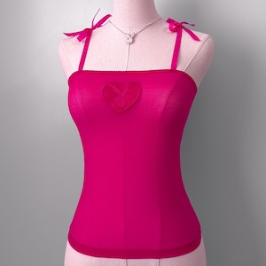 May include: A vibrant fuchsia camisole with a heart-shaped logo patch. The top features delicate bow-tied straps and matching trim. A silver necklace with a bunny pendant is also visible.