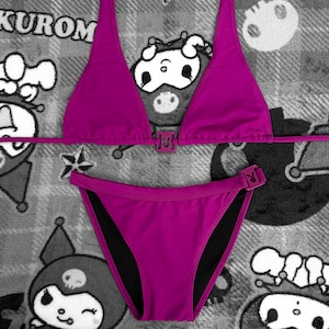 May include: A magenta bikini set with a triangle top and low-rise bottoms. The bikini features matching magenta buckles. The background is a gray and white patterned fabric with cartoon characters.