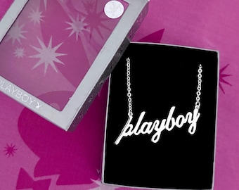 May include: A silver-colored necklace with the word "playboy" in cursive script, displayed in a black box. The box is open, revealing the necklace against a black background. The box is inside a silver box with a pink background.