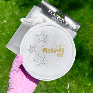 Puede incluir: Un estuche redondo y blanco para CD/DVD con el texto Playboy NYC en letras doradas y tres contornos de estrellas formados por pequeños diamantes de imitación plateados. El estuche está unido a una tarjeta negra y blanca con el texto Playboy CD/DVD Case.