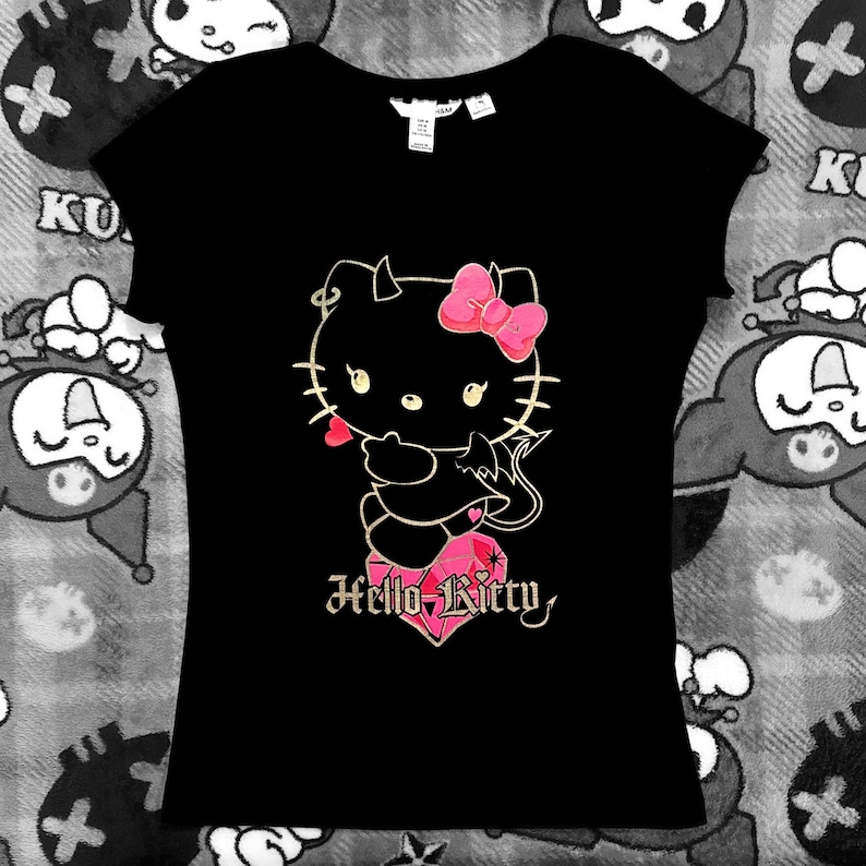 May include: Black t-shirt featuring a cartoon Hello Kitty character with devil horns, a pink bow, and a heart-shaped gem. The text "Hello Kitty" is below the character in a gothic font. The shirt has a crew neck and short sleeves.