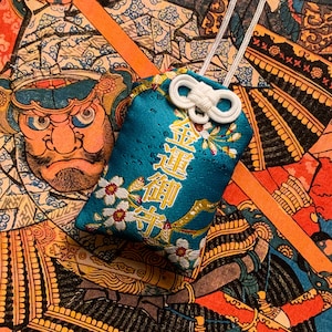 May include: A teal blue fabric charm with gold embroidery and white flowers. The charm has the Japanese characters "金運 御守" which translate to "Good Fortune Charm".