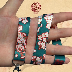 Peut inclure: Un cordon en tissu sarcelle orné d'un motif répétitif de fleurs de cerisier blanches et rose clair avec des contours rouges. Le cordon comprend un mousqueton pivotant en métal argenté à la base pour attacher des objets en toute sécurité.