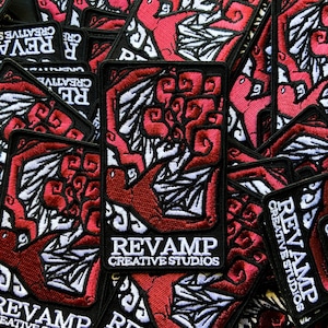 Op de afbeelding: Een close-up van verschillende rode en zwarte geborduurde patches met de tekst "REVAMP CREATIVE STUDIOS" in wit. De patches hebben een gestileerd ontwerp van een vogel met uitgespreide vleugels.