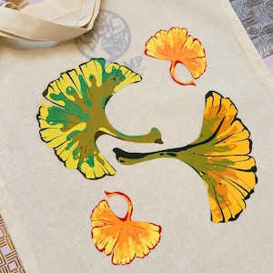 Può includere: Una borsa tote in tela beige con un design serigrafiato di quattro foglie di ginkgo. Due foglie grandi mostrano una miscela di tonalità gialle e verdi, mentre due foglie più piccole sono colorate in vivaci sfumature di arancione e giallo.