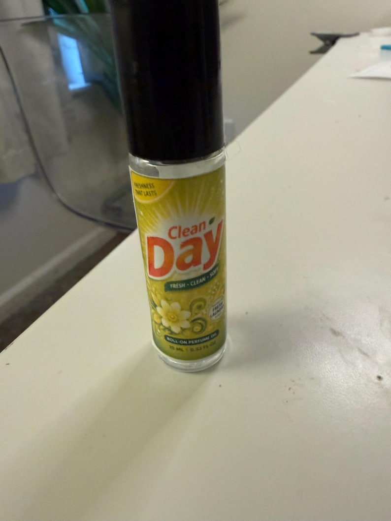 May include: A roll-on perfume bottle with a black cap and a yellow and green label. The label features the words "Clean Day" in red and white, along with floral graphics. The bottle contains 10 ml / 0.33 fl oz of perfume.