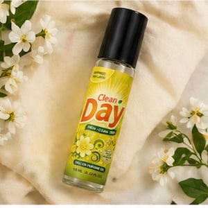 May include: A clear glass bottle of "Clean Day" perfume oil with a black cap. The label is yellow with orange and green text and a floral design. The bottle is surrounded by white flowers and green leaves.