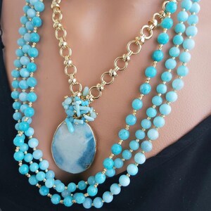 May include: A layered necklace with a large, oval, blue and white pendant. The pendant is surrounded by smaller blue stones. The necklace is made of gold chain and blue beads.
