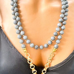 Puede incluir: Un collar de doble hebra con cuentas grises redondas y facetadas. La secci&oacute;n inferior incorpora una cadena de metal dorado con eslabones circulares, conectada a un colgante de piedra gris facetada en el centro del dise&ntilde;o.