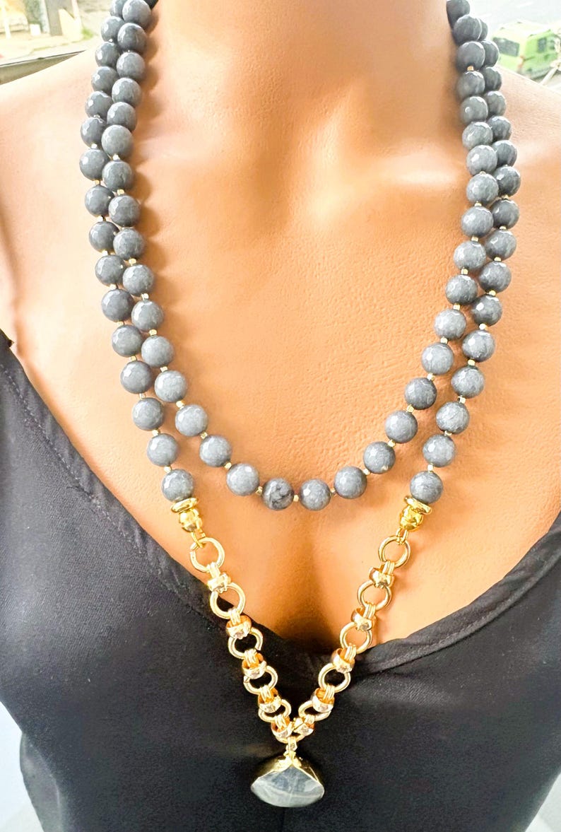 Puede incluir: Un collar de doble hebra con cuentas grises redondas y facetadas. La secci&oacute;n inferior incorpora una cadena de metal dorado con eslabones circulares, conectada a un colgante de piedra gris facetada en el centro del dise&ntilde;o.