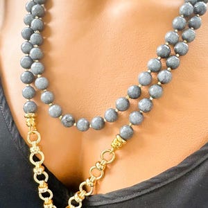 Puede incluir: Un collar de doble hebra con cuentas grises facetadas y detalles en metal dorado. El collar incluye una secci&oacute;n de cadena de eslabones gruesos en tono dorado que se conecta a un colgante de piedra gris, creando una apariencia de capas.
