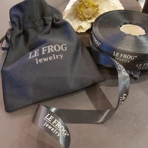 Puede incluir: Una bolsa negra con cord&oacute;n y un rollo de cinta de sat&eacute;n negro a juego, ambos con el texto impreso en blanco LE FROG Jewelry. La cinta est&aacute; parcialmente desenrollada, mostrando el nombre de la marca y el logotipo repetidos.