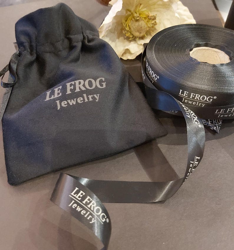 Puede incluir: Una bolsa negra con cord&oacute;n y un rollo de cinta de sat&eacute;n negro a juego, ambos con el texto impreso en blanco LE FROG Jewelry. La cinta est&aacute; parcialmente desenrollada, mostrando el nombre de la marca y el logotipo repetidos.