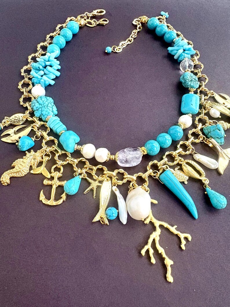 May include: A gold-toned necklace featuring turquoise beads, coral, and charms. The necklace includes a seahorse, anchor, fish, and coral branch pendants. Additional elements include white pearls and clear crystal beads.