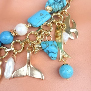 May include: A gold-toned chain necklace with turquoise, white, and clear beads. The necklace features gold charms of a mermaid tail, fish, and seashell. The turquoise beads are a mix of round and rectangular shapes.