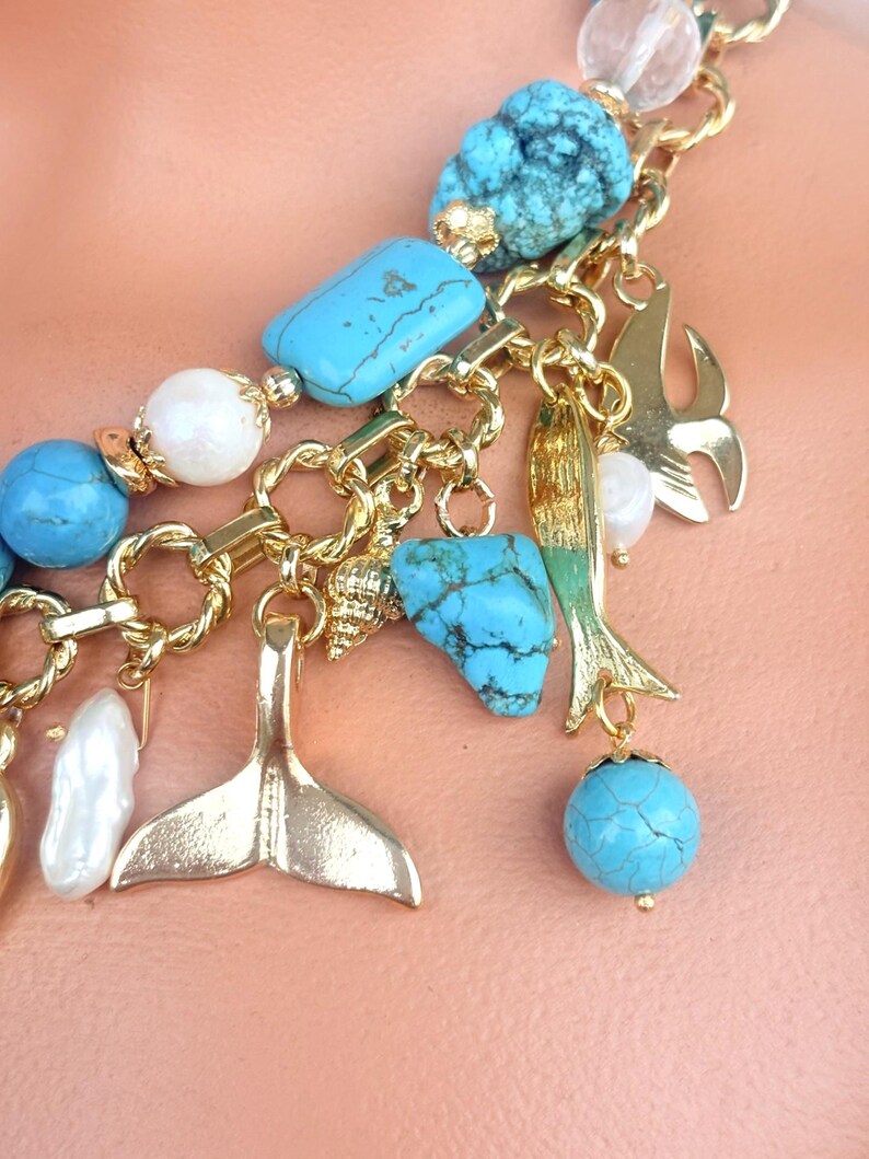 May include: A gold-toned chain necklace with turquoise, white, and clear beads. The necklace features gold charms of a mermaid tail, fish, and seashell. The turquoise beads are a mix of round and rectangular shapes.