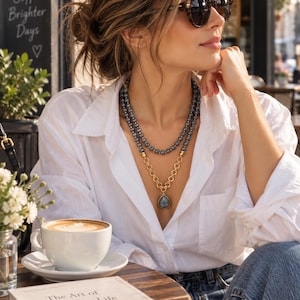 Puede incluir: Un collar de varias capas que presenta una hilera de cuentas de color gris oscuro y una cadena de tono dorado con un colgante en forma de l&aacute;grima. El collar se lleva sobre una camisa blanca de botones. La joya combina eslabones dorados.