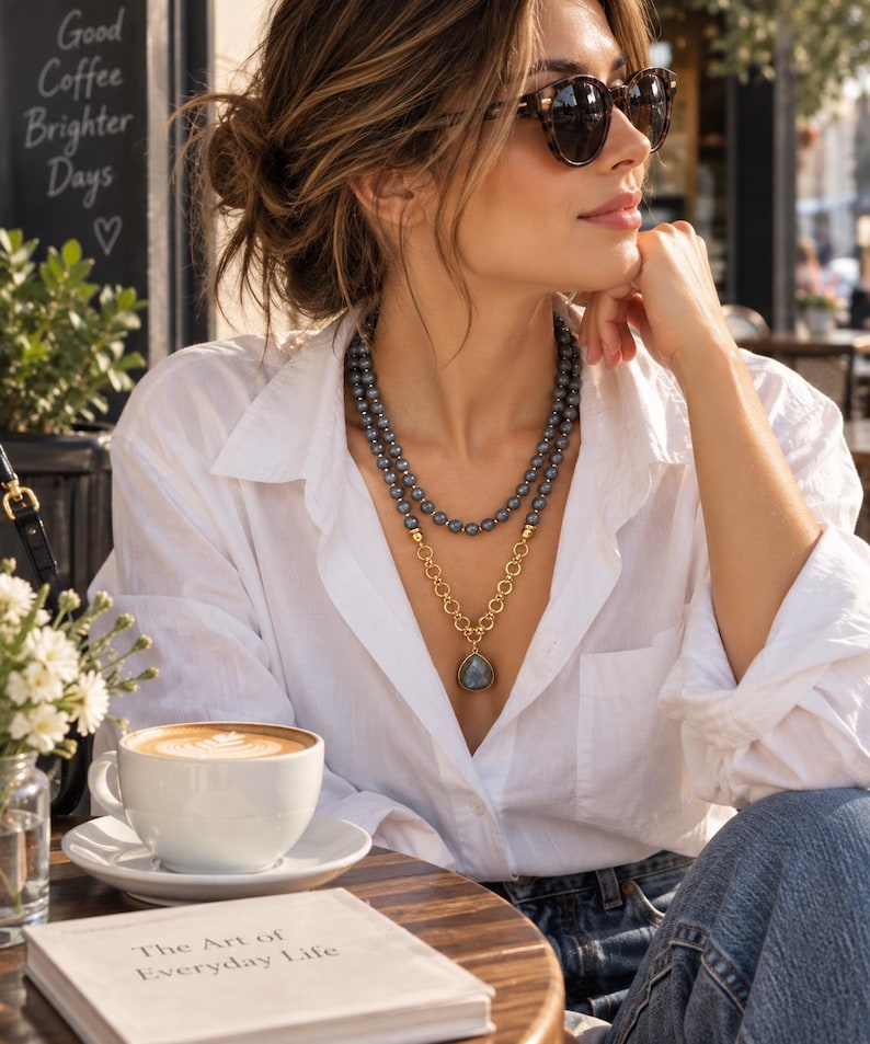 Puede incluir: Un collar de varias capas que presenta una hilera de cuentas de color gris oscuro y una cadena de tono dorado con un colgante en forma de l&aacute;grima. El collar se lleva sobre una camisa blanca de botones. La joya combina eslabones dorados.