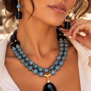 Puede incluir: Un collar de varias hebras con dos filas de cuentas azules moteadas, cuentas negras y un gran colgante ovalado negro con detalles dorados. Los pendientes a juego consisten en un poste dorado texturizado, una cuenta azul y un cilindro negro.